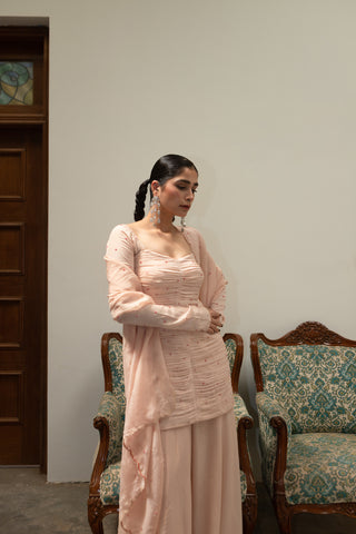 Side profile of baby pink ruched peplum set showing body-hugging fit, embellished sleeves, and layered sharara.