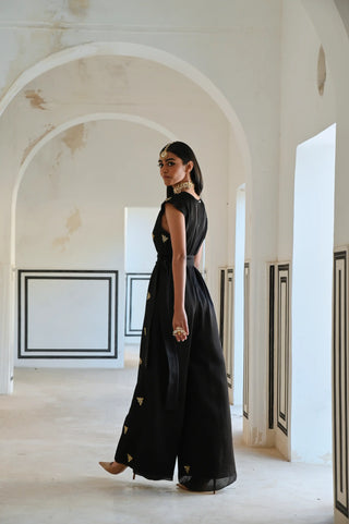A woman wearing a black sleeveless jumpsuit dress with gold embroidered details walks through a white arched corridor.
