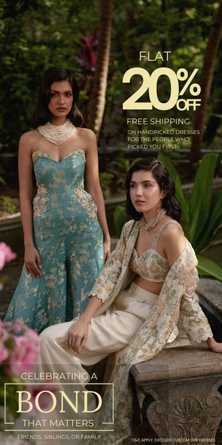 Two women modeling contemporary Indian ethnic wear jumpsuits in an outdoor garden setting. One wears a strapless teal-blue embroidered jumpsuit, and the other wears a cream and beige embroidered Jacket with Pant set. Perfect dress for cocktail parties, wedding reception and wedding welcome parties.