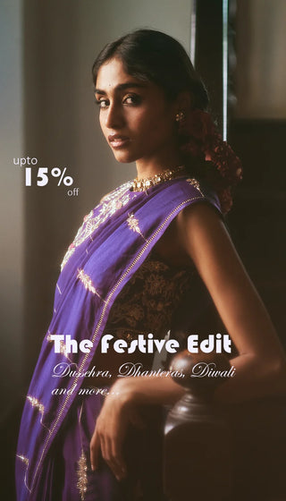 A woman wearing a purple sari with gold embroidery and a black embroidered blouse, posed in a dimly lit indoor setting. White promotional text overlays read 'upto 15% off' and 'The festive Edit' with smaller festive-themed copy. Dussehra, Dhanteras, Diwali and more.. 