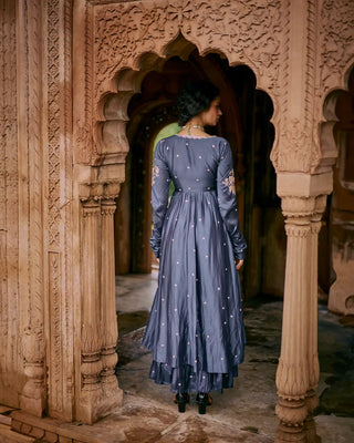A woman is back posing to show the anarkali with skirt look. It has hand-emboridery butti's all over the blue dress with churidar long sleeves. 