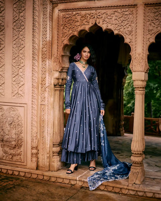 Woman in a blue anarkali dress standing in an ornate architectural setting. She is wearing an elegant modal silk anarkali featuring hand-embroidered zardozi and pita work buttis all over. It is paired with a hand-embroidered skirt and our signature Roop flip bird printed hand-embroidered dupatta. Designed by Paulmi and Harsh. Shop at Leela Lane