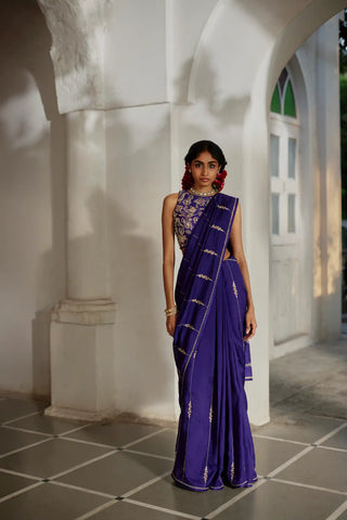 A women in a solid purple saree adorned with exquisite hand-embroidered lines and delicate floral accents. Paired with a matching blouse fully embellished with aari and zardozi embroidery, and finished with elegant hand-tucked tassel details for a sophisticated, artisanal look.