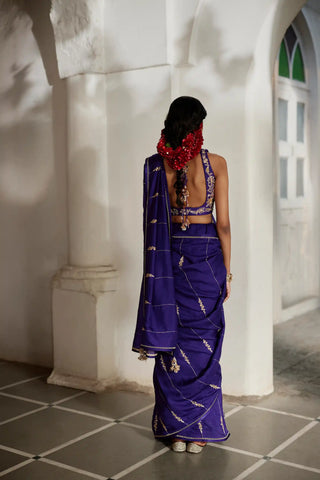 A woman viewed from behind wearing a deep purple saree with gold zari embroidery, standing in a white-walled corridor with tiled flooring.