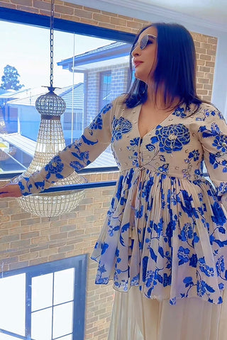 Woman wearing a blue floral embroided peplum with palazzo dress standing by a window with a chandelier.  A perfect dress for festive forward glam, celebrations, family gatherings and ethnic chic parties.