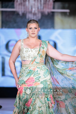 Model in rich green and ivory floral printed pre-draped chiffon ruffle saree featuring elegant prints and tiered lace detailing that adds a fluid movement. It is paired beautifully hand-embroidered blouse at fashion runway.