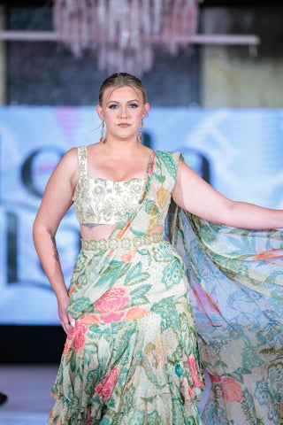 Model in rich green and ivory floral printed pre-draped chiffon ruffle saree featuring elegant prints and tiered lace detailing that adds a fluid movement. It is paired beautifully hand-embroidered blouse at fashion runway.