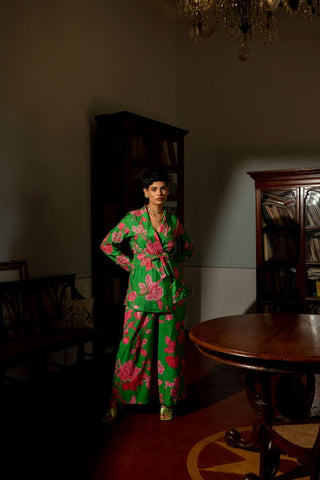 A woman stands in a dimly lit room wearing a bright green two-piece outfit with a bold pink floral print. The set includes a long-sleeve belted jacket and matching wide-leg pants. Perfect for parties, casual outings, brunch parties, formals or summer day out with family.