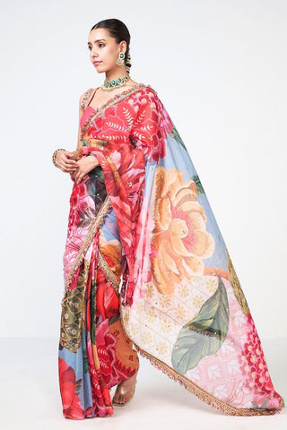 A woman modeling a vibrant floral saree with a red, pink, and light blue print, featuring a gold embroidered border and a large peach-colored floral motif on the drape, set against a plain white studio background.