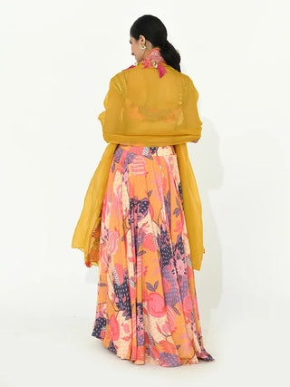 A woman is shown from behind wearing a vibrant, multicolored floral lehenga printed skirt with a solid mustard yellow sheer shawl draped over her shoulders, set against a plain white studio background.